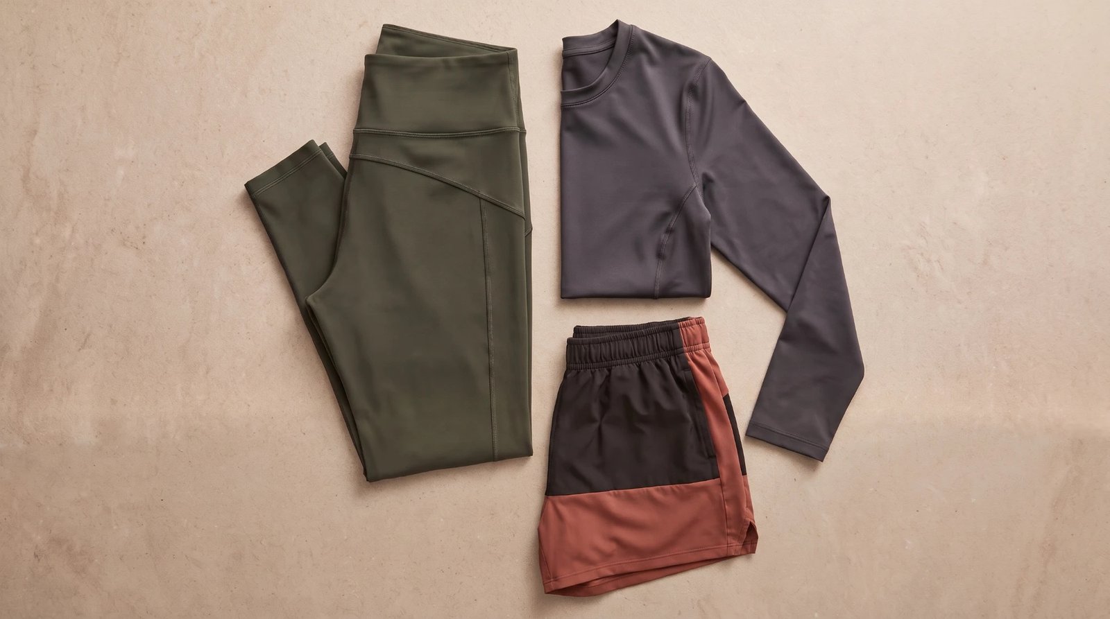Activewear flat lay on a cream backdrop: olive leggings, a gray long-sleeve top, and terracotta shorts